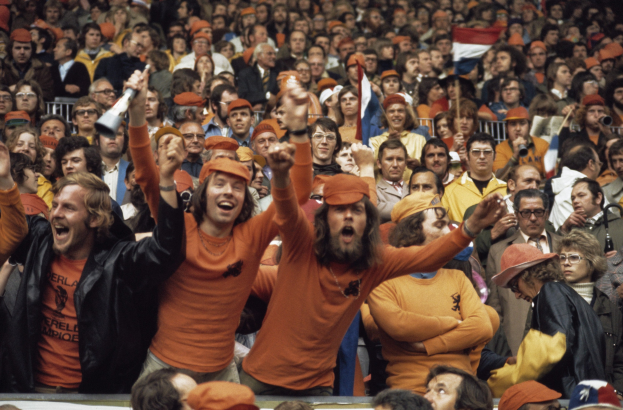Eine große Menge in orangen T-Shirts und Hüten jubelt in einem Stadion, einige halten Fahnen und eine Person hat ein Megafon, mit Geländern im Hintergrund.