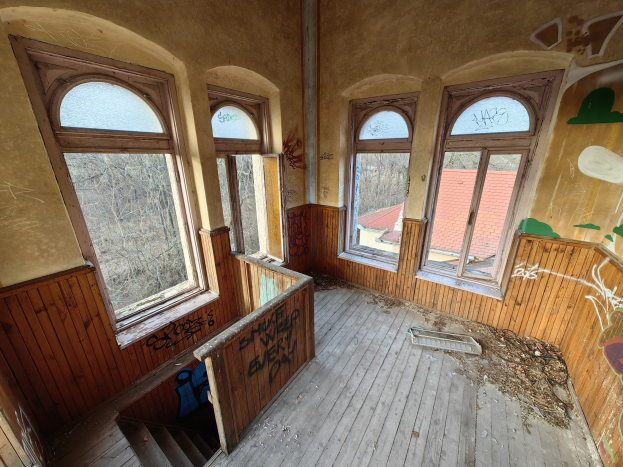 Verlassener Gebäuderaum mit Graffiti an den Wänden und Fenstern, Holzboden, Treppe, verstreuter Trümmer und Bäumen im Hintergrund.