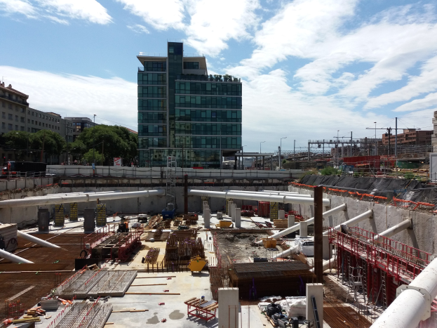 Baustelle mit einem großen Gebäude im Hintergrund, umgeben von Bäumen, Pfählen, Lampen und anderen Gegenständen, unter einem bewölkten Himmel, als Europäischer Union Hauptsitz identifiziert.