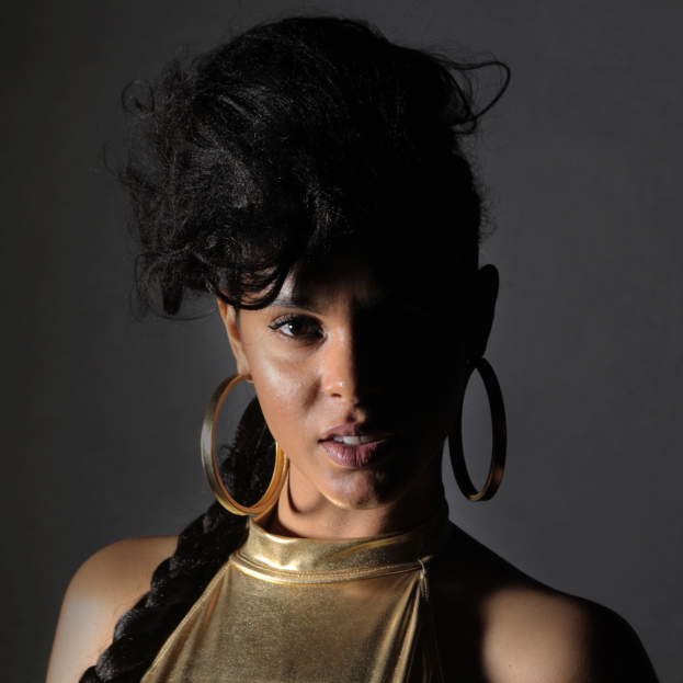 Eine Frau in einem goldenen Kleid und großen Ohrringen steht selbstbewusst vor einer Wand, ihre Haare sind ordentlich frisiert, mit dezentem Make-up und einem entschlossenen Gesichtsausdruck.