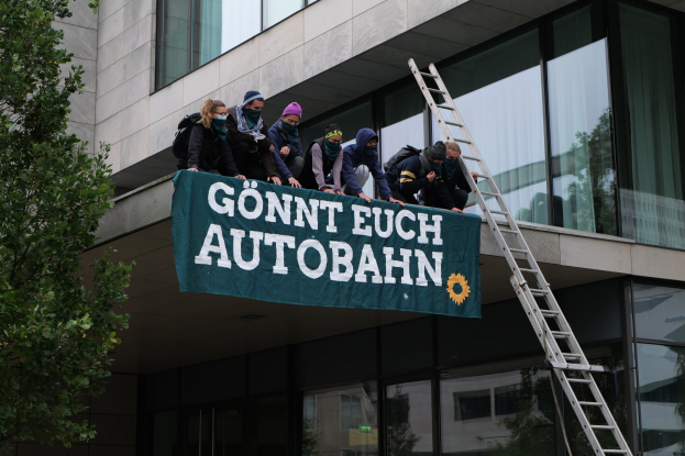 Eine Gruppe von Menschen steht auf dem Dach eines Gebäudes und hält ein Transparent in die Höhe, mit einem Baum links und Glasfenstern im Hintergrund, sie scheinen für die deutsche Regierung zu protestieren.