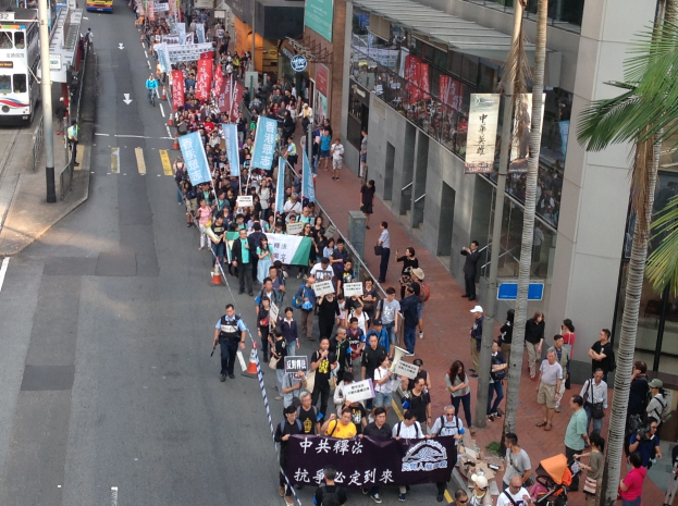 Eine große Gruppe von Menschen marschiert auf einer Stadtstraße mit Transparenten und Schildern, mit Bäumen, Gebäuden, Fahrzeugen und Straßenschildern im Hintergrund.