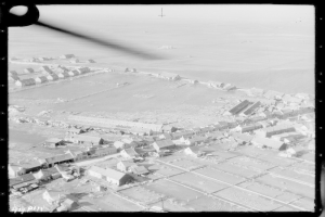 Eine Luftaufnahme einer Kleinstadt mit zahlreichen Häusern und umliegender Landschaft, dargestellt in Schwarz-Weiß.