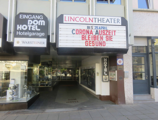 Außenansicht des Lincoln Theaters in Berlin, Deutschland, mit Glasfenstern und -türen sowie einer Tafel mit Text und einer belebten Stadtkulisse im Inneren.