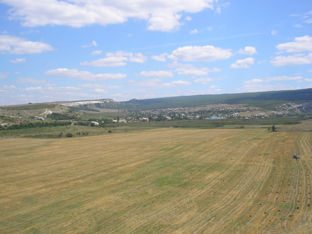 Ein Traktor, der auf einem kultivierten Feld umgeben von Bäumen, Gebäuden, Hügeln und einer bewölkten Himmel operiert.