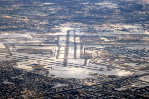Luftaufnahme eines städtischen Flughafens umgeben von schneebedeckten Straßen, Gebäuden, Bäumen und einer weiten winterlichen Landschaft.
