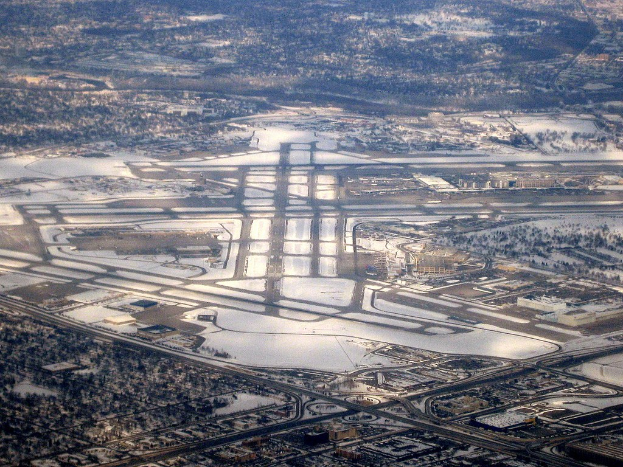 Luftaufnahme eines städtischen Flughafens umgeben von schneebedeckten Straßen, Gebäuden, Bäumen und einer weiten winterlichen Landschaft.