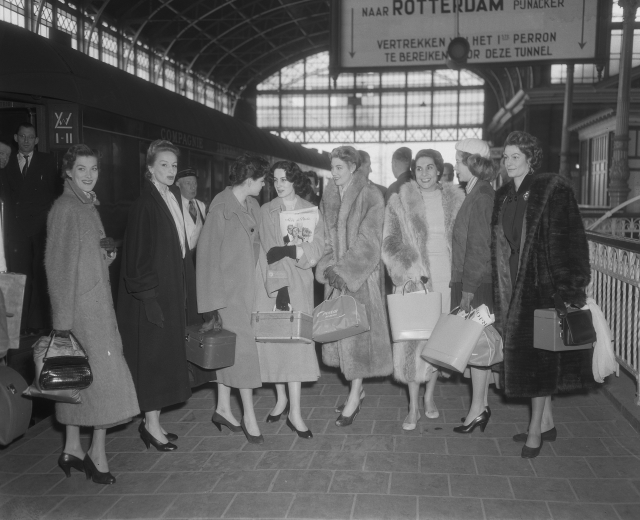 Schwarz-weißes Bild einer Gruppe von Frauen, die an einem Bahnhof stehen, jede hält eine Tasche, mit einem Zug auf der linken Seite und Geländern und einer Tafel auf der rechten Seite.