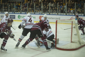 Eine Gruppe von Menschen, die Hockey auf einem Eisplatz spielen, tragen Sportkleidung und halten Stöcke, mit einem Torpfosten auf der rechten Seite, Tafeln mit Text, Pfosten und Zuschauern im Hintergrund.