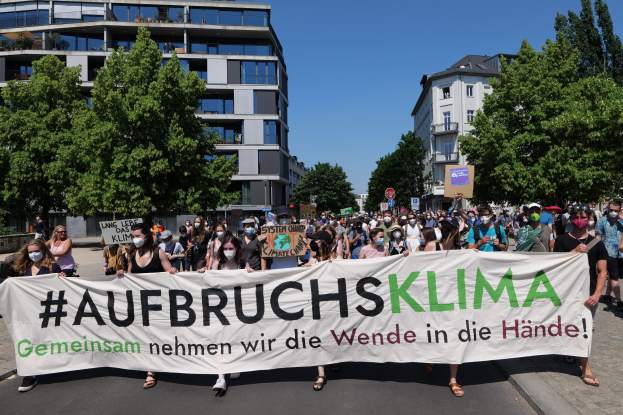 Gruppe von Menschen mit Masken, die ein Banner mit der Aufschrift 'Aufbruchsklima' vor einem Gebäude mit Bäumen und einem klaren Himmel halten.