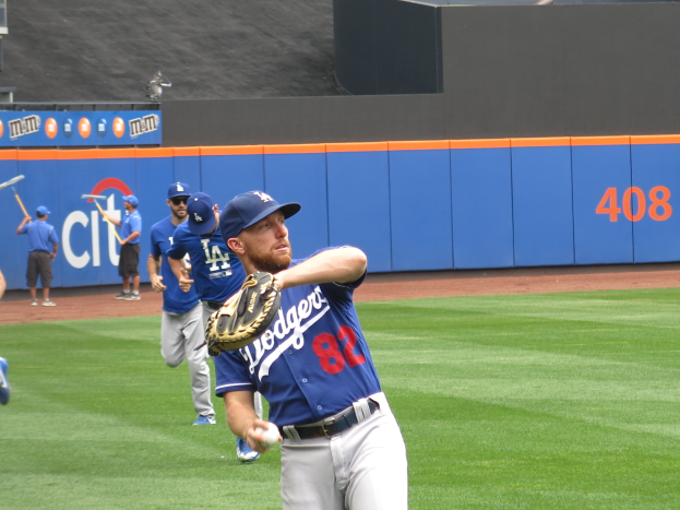 Ein Baseballspieler in einem blauen T-Shirt, weißen Hosen und einer blauen Mütze wirft einen Ball auf einem Feld, trägt einen braunen Baseballhandschuh und hat andere Spieler im Hintergrund, einige halten Baseballschläger.