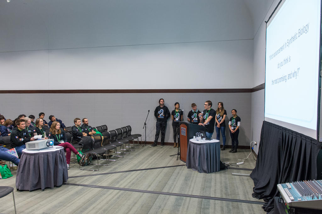 Eine Person hält ein Mikrofon und spricht zu einem Publikum in einem Bürosetting, wobei Teammitglieder Namensschilder tragen und eine Präsentation hinter ihnen angezeigt wird.