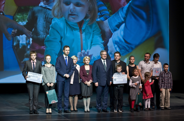 Gruppe von Menschen auf einer Buhne mit Taschen und Urkunden, im Hintergrund ein Bildschirm mit einem Kind und anderen.