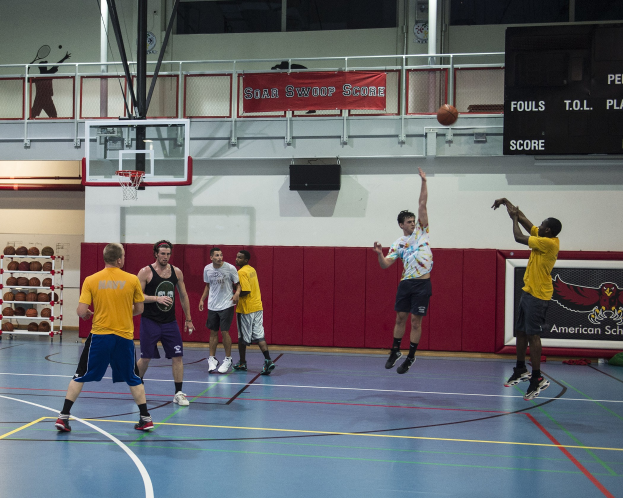 Gruppe von Männern, die Basketball in einer Turnhalle mit einem Scoreboard auf der rechten Seite und einem Basketballkorb in der Mitte spielen.