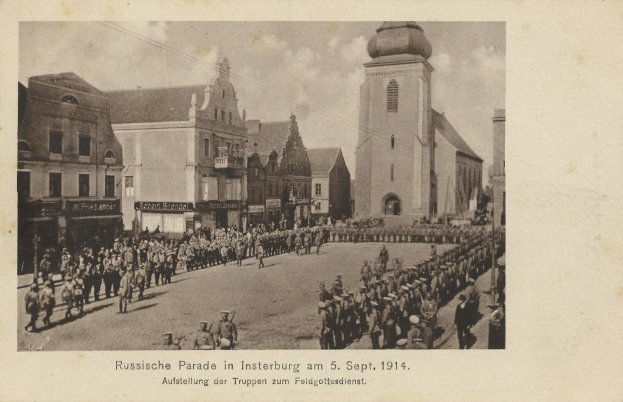 Ein Schwarz-Weiß-Foto eines Umzugs in Insterburg am 5. September 1914 mit vielen Menschen, Gebäuden und bewölktem Himmel sowie Text am unteren Rand.