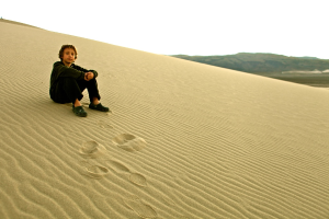 Ein Junge sitzt auf Sand mit Bergen im Hintergrund und Himmel darüber.