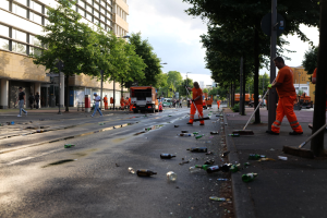 Eine Gruppe von Menschen in orangefarbenen Uniformen, die Müll auf einer Straße mit Flaschen und Schutt säubert, mit Bäumen, Pfählen und Fahrzeugen im Hintergrund unter einem bewölkten Himmel.