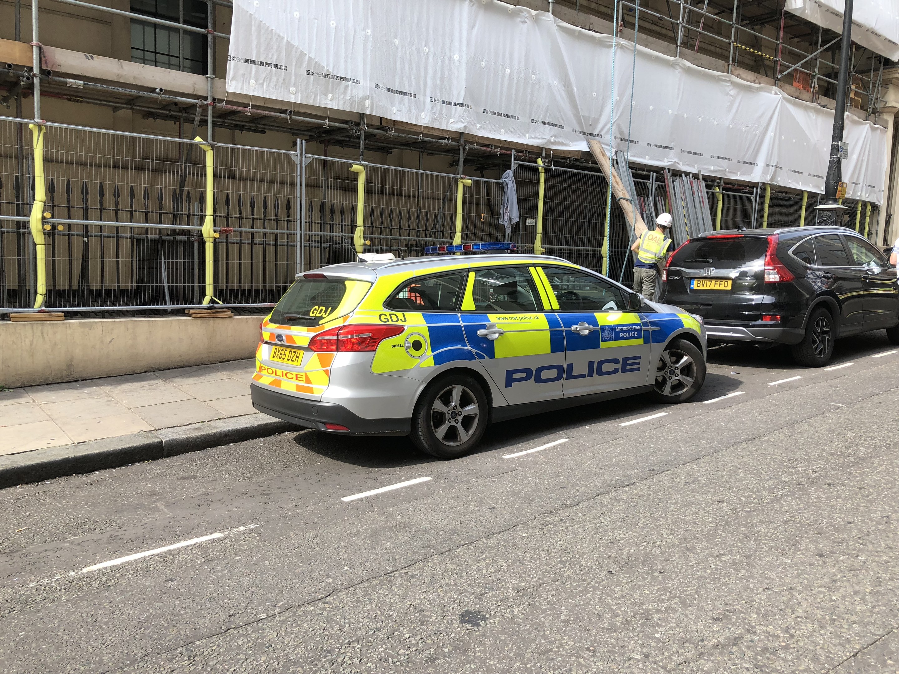 Polizeiauto neben einem Gebäude geparkt, mit zwei Helmen tragenden Personen und einer weißen Abdeckung im Hintergrund, was auf eine Londoner Kriminalermittlung hindeutet.