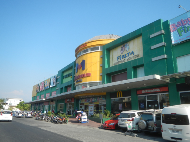 Ein Einkaufszentrum mit parkenden Autos, umgeben von Gebäuden, Werbetafeln, Geschäften, Motorrädern, Pflanzen, Bäumen, Straßenlaternen und einer bewölkten Himmel.