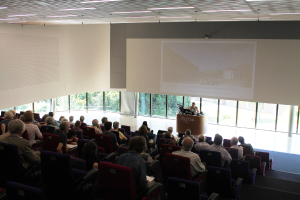 Ein Seminarraum in einer Universität mit vielen Sitzenden, die einem Präsentator an einem Tisch gegenübersitzen, der eine Präsentation zeigt, mit Glasfenstern, die Bäume und Gebäude im Hintergrund zeigen.