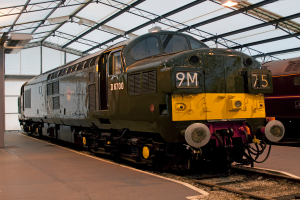 Ein Zug befindet sich auf den Schienen neben einem Bahnsteig mit einem Mast auf dem Dach, der Metallstangen mit Lampen trägt.