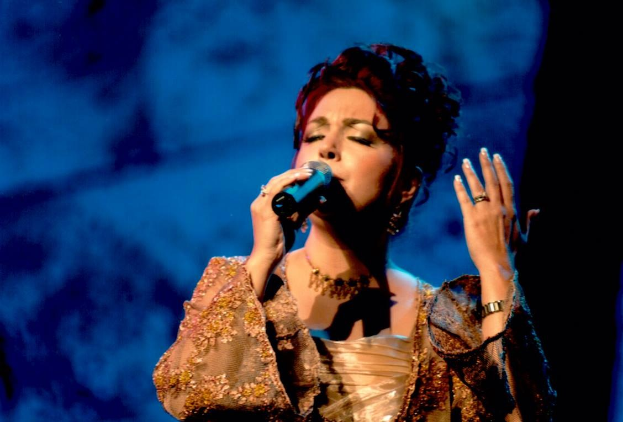 Frau in goldenem Kleid singend ins Mikrofon auf der Bühne bei der Eurovision mit blau-schwarzem Hintergrund.