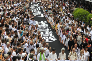 Eine große Gruppe von Menschen marschiert auf einer Straße, hält Protestschilder und Banner hoch und hat Grünzeug auf der rechten Seite und ein Gebäude im Hintergrund.