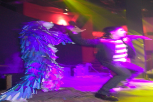 Leute tanzen auf der Bühne, wobei eine Person links ein elegantes Kleid trägt und Scheinwerfer oben sichtbar sind.