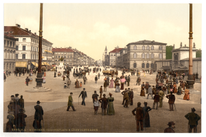 Ein altes Schwarz-Weiß-Foto einer belebten Münchner Stadtstraße aus den frühen 1900er-Jahren, das Fußgänger, einige mit Schirmen, einen Pferdewagen im Vordergrund, Gebäude, Bäume und Pfähle im Hintergrund zeigt, mit Text am unteren Bildrand.