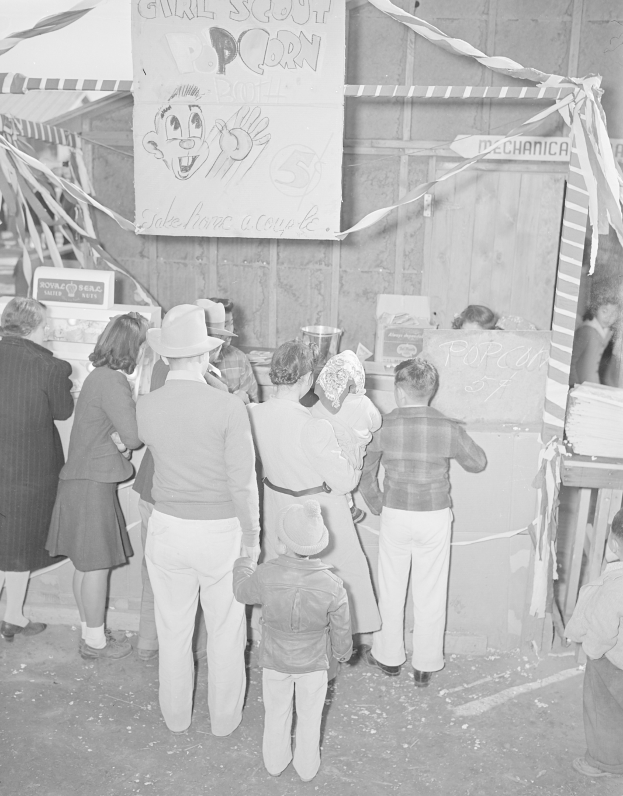 Schwarzes und weißes Foto einer Gruppe von Menschen, die vor einem Karnevalsstand mit Bändern und einer Schautafel stehen.