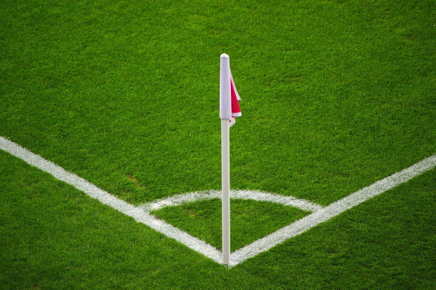 Ein Fussballfeld mit gruener Gras und weissen Begrenzungslinien, mit einem weissen Pfosten mit einer roten und weissen Flagge, die den Elfmeterpunkt markiert.