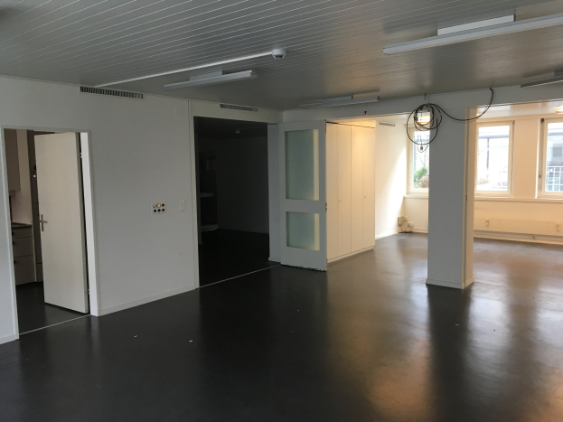 Leerer Büroraum mit schwarzen Böden, weißen Wänden, elektrischem Licht, Kabeln, Türen, Schränken, Fenstern und einem Außenblick auf Gebäude und Bäume in Amsterdam.