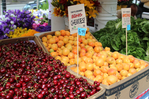 Ein Bauernmarktstand mit Kisten voller Kirschen, Orangen und Blattgemüse, Schilder mit Text und Menschen, Fahrzeuge, Pfosten und Gebäude im Hintergrund.