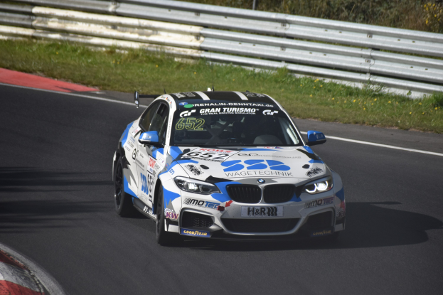 Ein BMW M4 DTM-Rennwagen mit Text und Zahlen auf der Karosserie, der auf einer Rennstrecke fährt, umgeben von Gras, einem Zaun und Pflanzen, mit sichtbarem Rad.