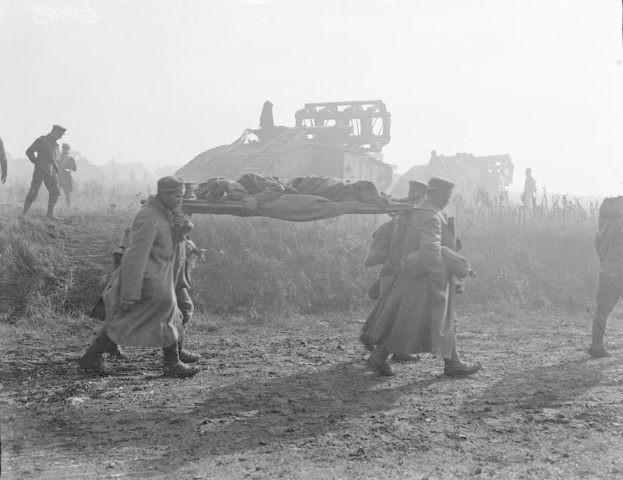 Eine Gruppe von Männern trägt eine verletzte Person auf einer Trage über einen Schotterweg, umgeben von Pflanzen und einem Tank im Hintergrund, dargestellt in Schwarz-Weiß.