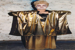 Eine Frau in einem goldenen Kimono und einem schwarzen und goldenen Kleid steht selbstbewusst vor einer weißen Backsteinwand.