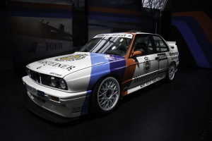Ein BMW E30 M3 Motorsport-Fahrzeug wird auf dem Boden mit einem Rad in der Mitte des Bildes gezeigt, umgeben von Bannern mit Text und Bildern im Hintergrund.