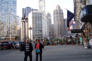 Eine belebte Straßenszene mit Menschen, Blumenkübeln, Autos, Straßenlaternen, Gebäuden, Geschäften und Fahnen auf der rechten Seite.