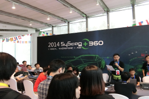 Ein Mann hält ein Mikrofon und spricht zu einer Gruppe, die an einem Tisch mit Laptops und elektronischen Geräten sitzt, unter einem Banner mit einem Design, Flaggen und Lichtern, in einem Raum mit Glasfenstern.