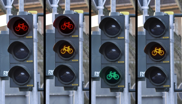 Ampel mit Fahrradsymbol, an einem Pfahl mit zusätzlichen Pfählen und einem Gebäude im Hintergrund angebracht.