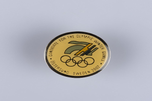 Runder Abzeichenaufkleber mit schwarzem Hintergrund und goldenem Olympische Winterspiele-Logo, mit fetter weißer Schrift auf einer weißen Oberfläche.