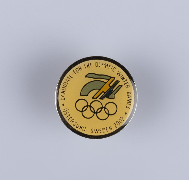 Runder Abzeichenaufkleber mit schwarzem Hintergrund und goldenem Olympische Winterspiele-Logo, mit fetter weißer Schrift auf einer weißen Oberfläche.