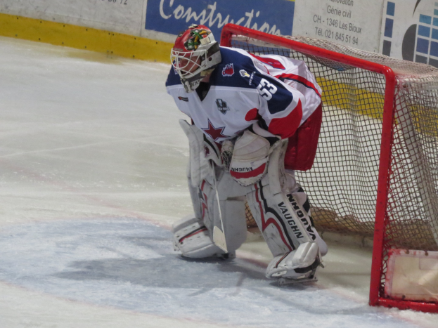 Torwart in voller Eishockey-Ausrüstung in einem Netz auf einem Eisstadion, mit einer textbedeckten Wand im Hintergrund.