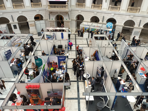 Eine große Gruppe von Menschen auf einer internationalen Messe mit Ständen, Bannern und einem Gebäude mit Bögen und Säulen im Hintergrund.