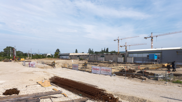 Baustelle mit verschiedenen Materialien, Geräten, Gebäuden, Kränen, Bäumen und wolkenverhangenem Himmel.
