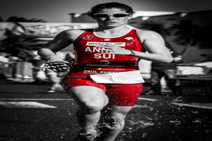 Eine Frau in einem roten Triathlonanzug läuft durch flaches Wasser, umgeben von einigen Menschen, Bäumen und einem Banner im Hintergrund, dargestellt in Schwarz-Weiß.