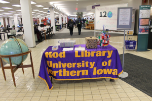 Eine Expo-Szene mit einem Tisch, auf dem verschiedene Landesflaggen, eine Kiste, Papiere und eine Fahne sowie links daneben eine Weltkugel und rechts ein Anschlagbrett mit einigen Menschen in der N√§he und mehreren Tischen und Stühlen im Hintergrund zu sehen sind.