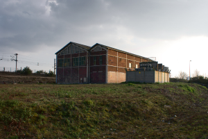 Eine Szene mit Schuppen, Transformatoren, elektrischen Masten mit Kabeln, Zäunen, einer Straßenlaterne, Bäumen und einem bewölkten Himmel.