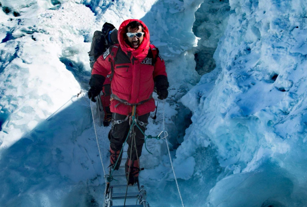 Ein Mann in einer roten Jacke, mit Skibrille, Handschuhen und einem Rucksack klettert einen schneebedeckten Berg hoch, gesichert mit Seilen und Equipment.