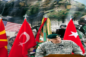 Ein Soldat steht an einem Podium mit einem Mikrofon, mit verschiedenen Flaggen und einem Plakat von Militärpersonal im Hintergrund.